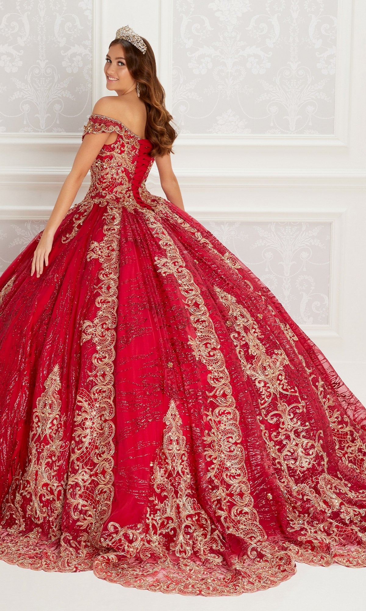 A woman wears the Quinceanera Dress PR22148 By Princesa, a red off-the-shoulder gown with gold lace embroidery and beaded accents, standing indoors against a white ornate wall and smiling over her shoulder.